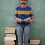 Schoolboy sitting on books stack and reading book